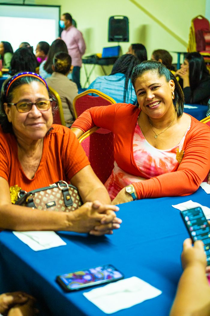 Smiling women enjoying a community gathering indoors, surrounded by people.