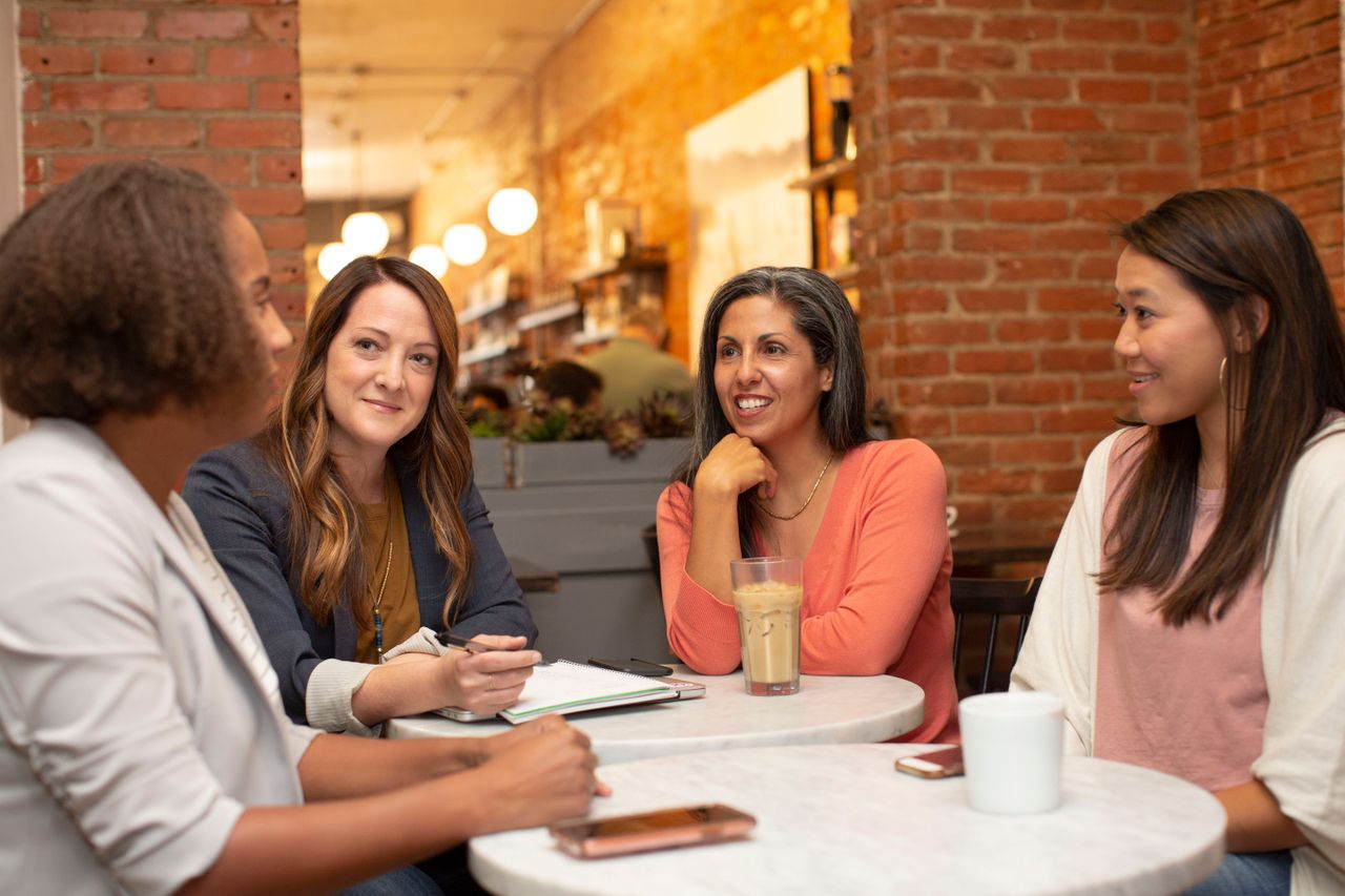 Four diverse women engage in a friendly discussion at a cafe, creating a warm and collaborative atmosphere.